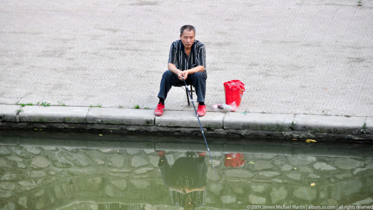 Man seated beside a stone-lined urban canal in Xi’an, fishing with a rod as his reflection appears in the still water, with a red bag and bright shoes beside him.