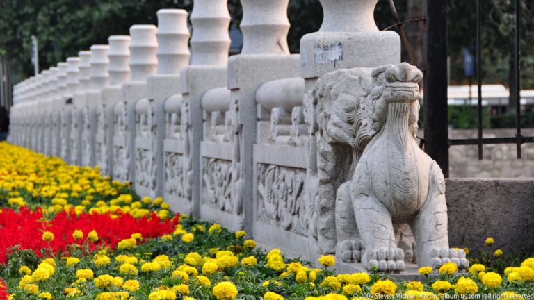 Carved Chinese guardian lion at the end of a stone balustrade in Xi’an, with matching stone posts receding behind and red and yellow flower beds below.