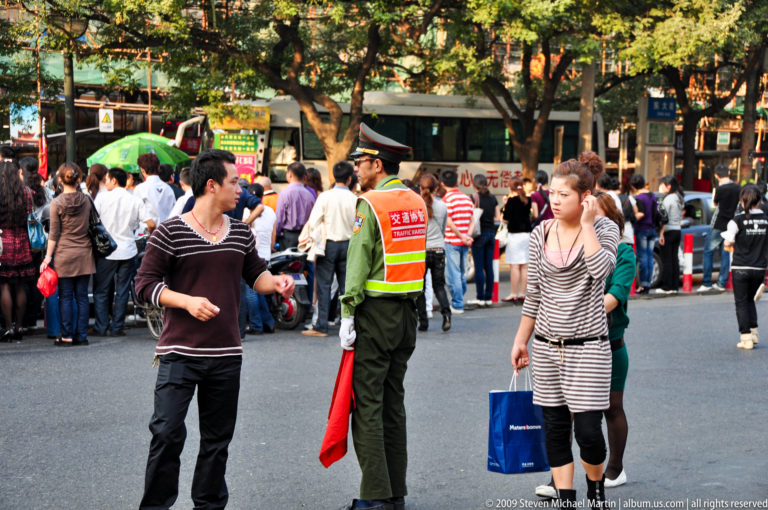 Traffic warden in reflective vest holding a red flag stands among pedestrians crossing a busy commercial street in Xi’an.