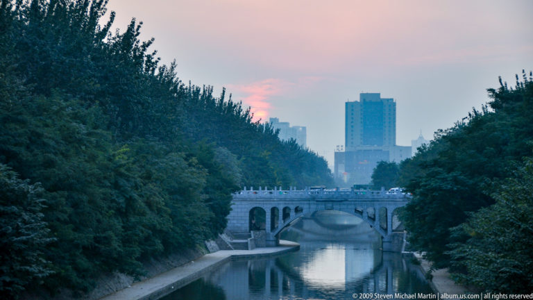 Tree-lined canal in Xi’an with stone embankments and a small arched bridge, with modern high-rise buildings in the distance.