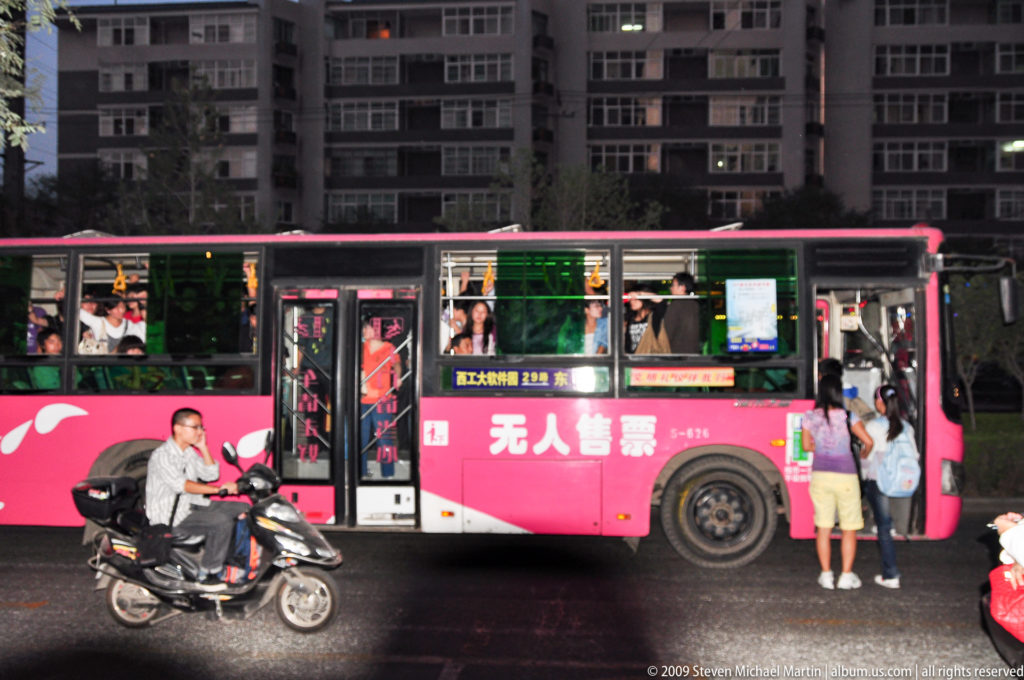 Pink city bus with Chinese advertising stopped on a Xi’an street beside a scooter rider and pedestrians, with apartment buildings in the background.
