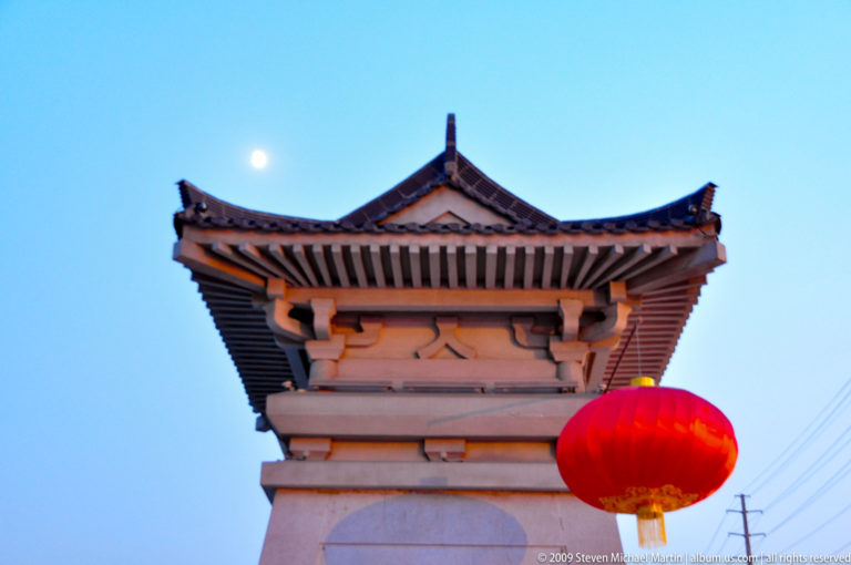Stone gatepost with small tiled roof and red lantern in Xi’an, silhouetted against a pale evening sky with the moon visible.