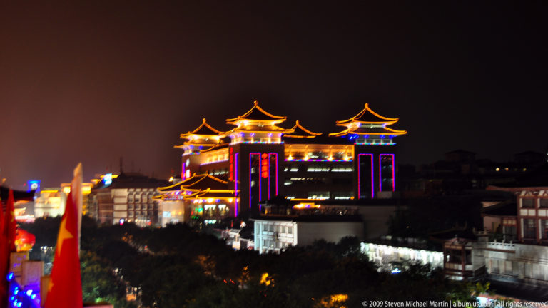 Night view of a large modern commercial complex in Xi’an with traditional pavilion roofs outlined in blue, magenta, and gold lighting, seen from above the surrounding dark city.