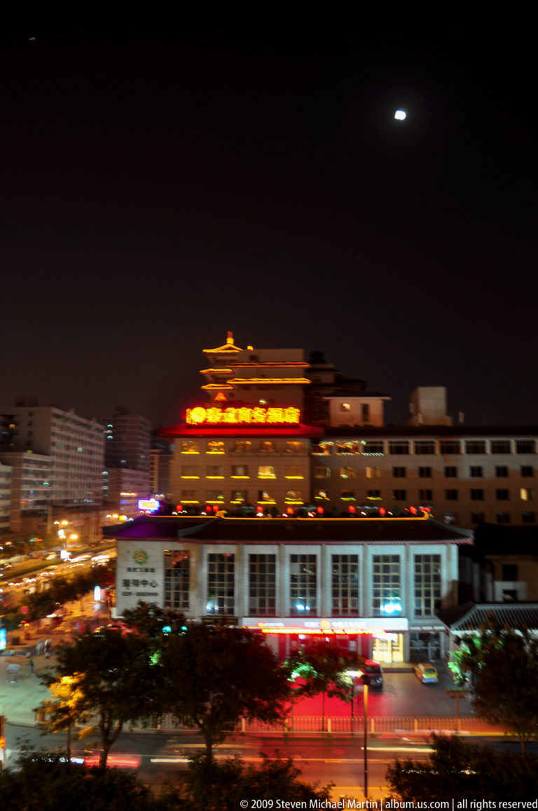 Nighttime cityscape in central Xi’an with a brightly lit multi-story commercial building topped by a traditional-style Chinese rooftop pavilion, framed by darker modern mid-rise structures.