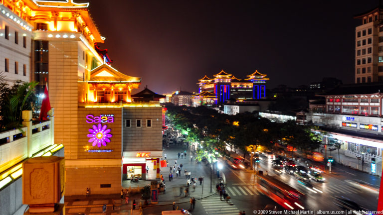 Elevated nighttime view of a busy Xi’an commercial street with traffic, pedestrians, traditional-style roofs, and illuminated signs including KFC and Sasa.