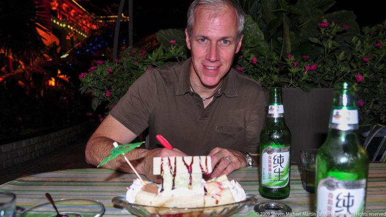 Male traveler smiling at a night restaurant table in Xi’an with a decorated dessert and green bottles of Chinese beer labeled 纯生.