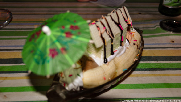 Boat-shaped dish with chocolate-drizzled cake slices, whipped cream, sprinkles, and a green paper parasol.