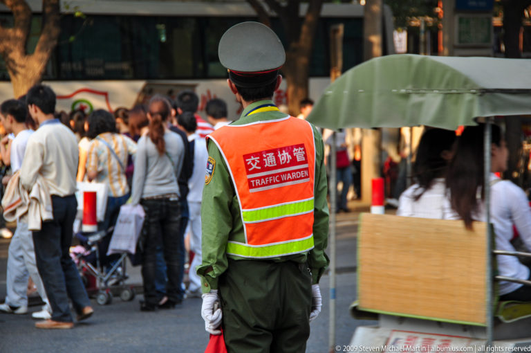 Rear view of a uniformed traffic warden in a high-visibility vest marked 交通协管 and TRAFFIC WARDEN, monitoring a dense pedestrian crowd in central Xi’an.
