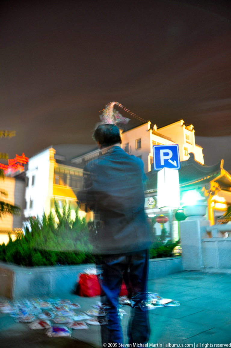 Blurred figure seen from behind at night in a brightly lit Xi’an street, holding a kite-like illuminated object beneath traditional-style roofs and a blue parking sign.