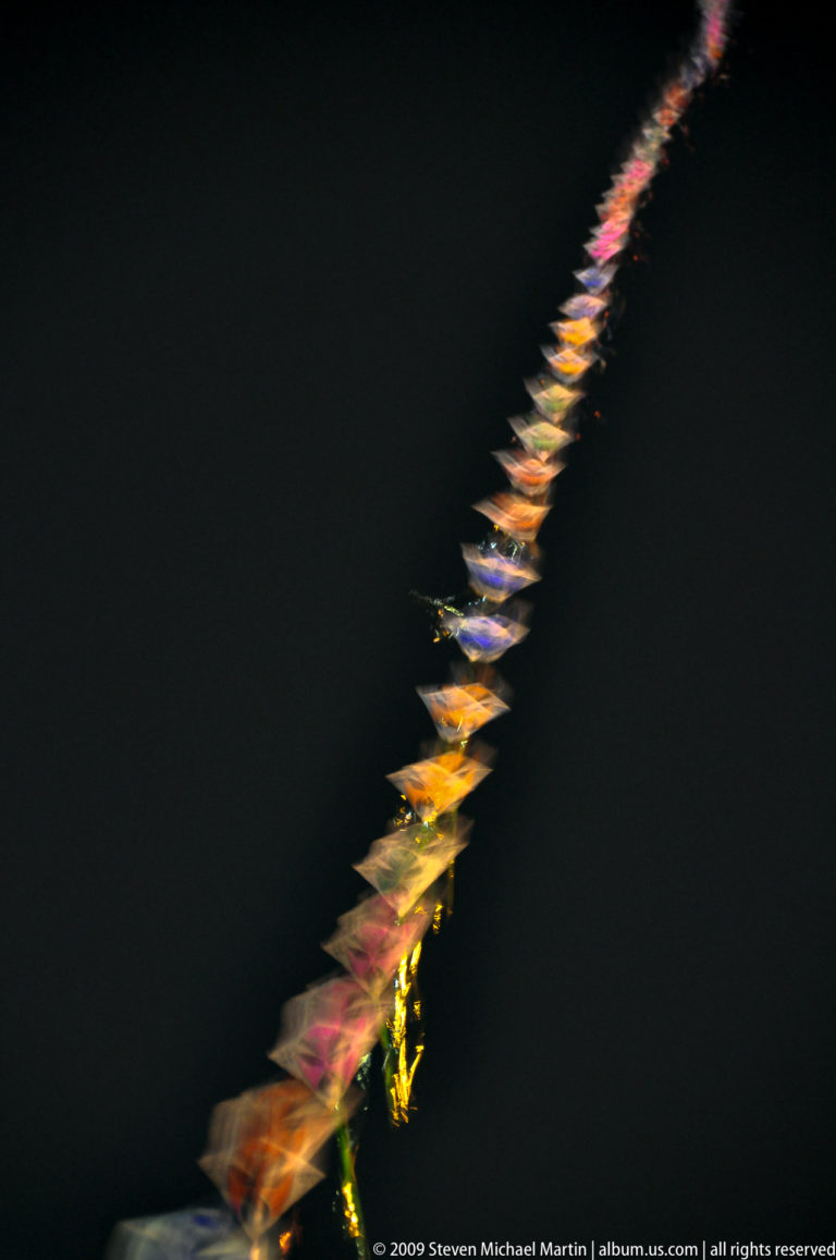 A long diagonal train of small illuminated kite- or pinwheel-like forms rises into a dark night sky.