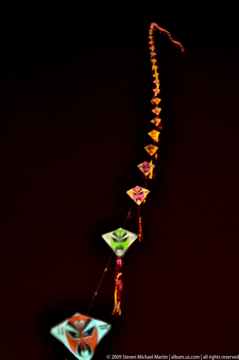 Vertical train of small illuminated diamond-shaped kites or wind toys receding upward into the night sky.