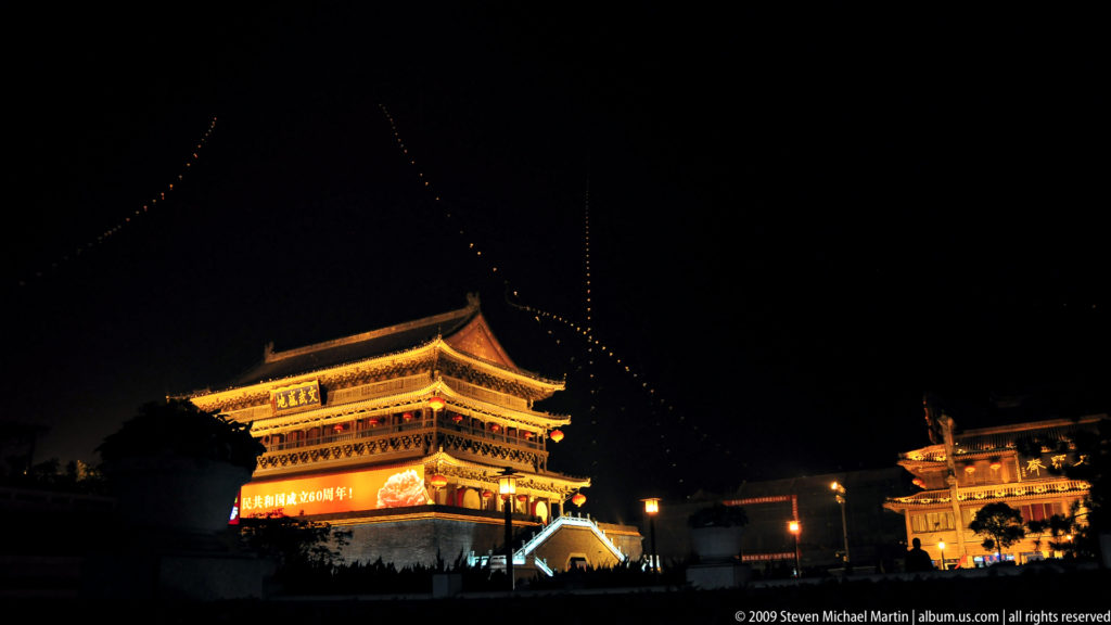 Distant night view of the illuminated Drum Tower of Xi’an rising above a dark public square, its layered roofs and high brick base clearly outlined by lights.