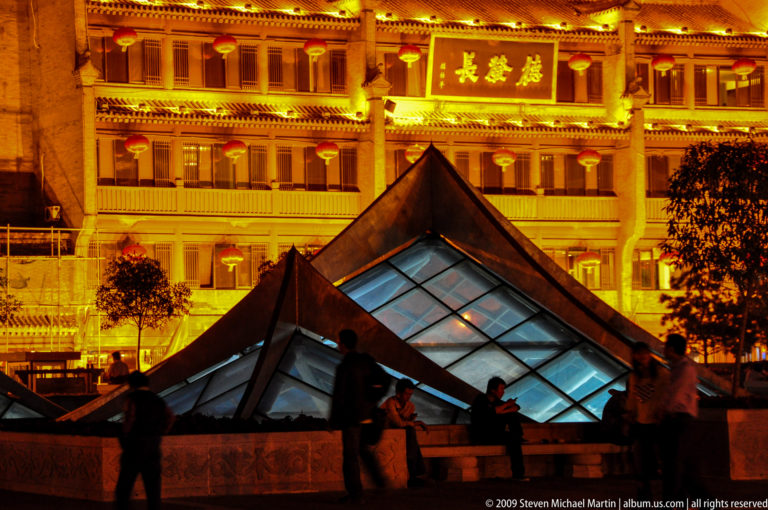 Night view of two low illuminated glass pyramid skylights in a Xi’an public plaza, with visitors in front of a warmly lit traditional-style commercial building in the background.