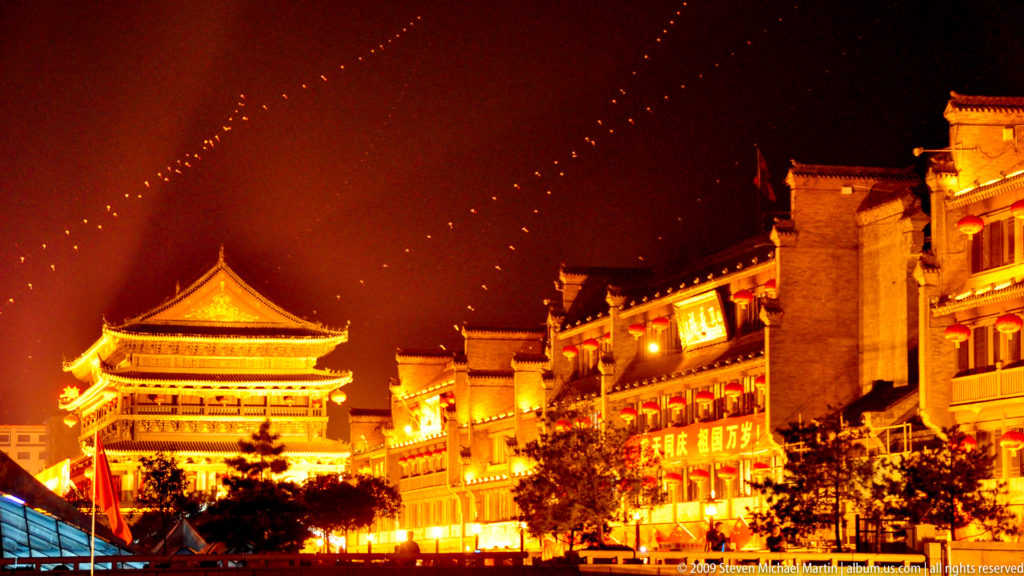 Night view of a multi-eaved ceremonial tower beside a brightly lit traditional-style commercial street in central Xi’an.
