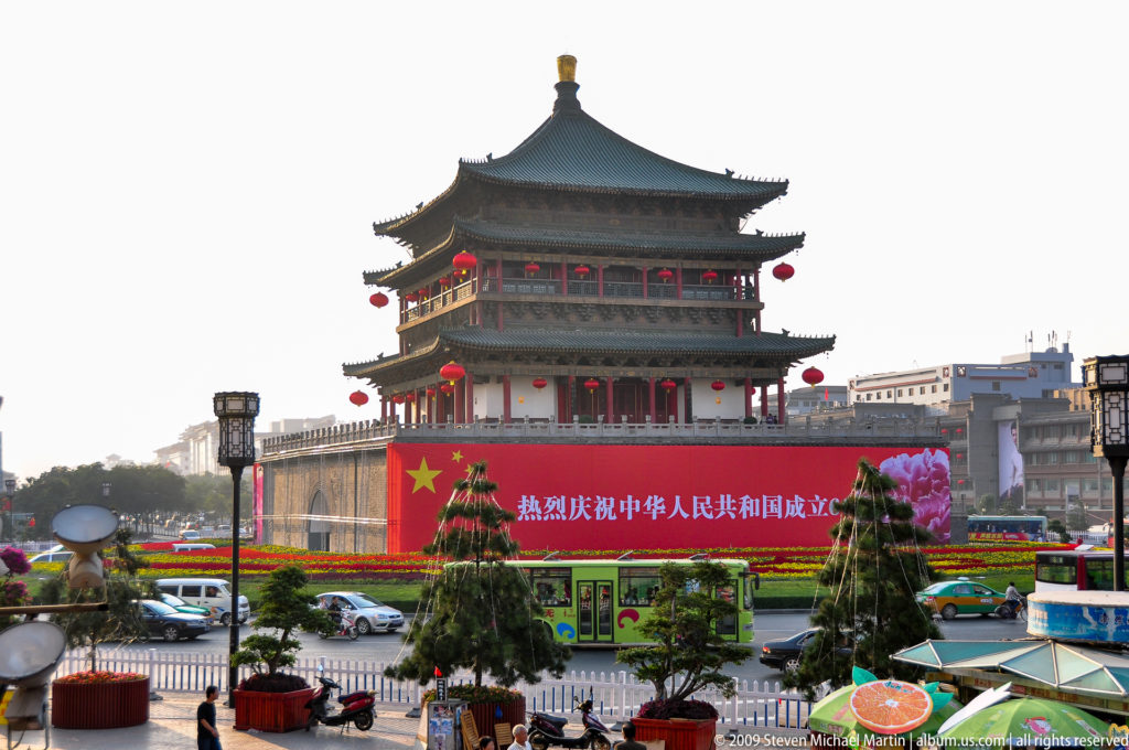 Bell Tower of Xi’an on a high masonry base with a large 60th-anniversary banner, surrounded by cars, buses, and flower displays in a central traffic circle.