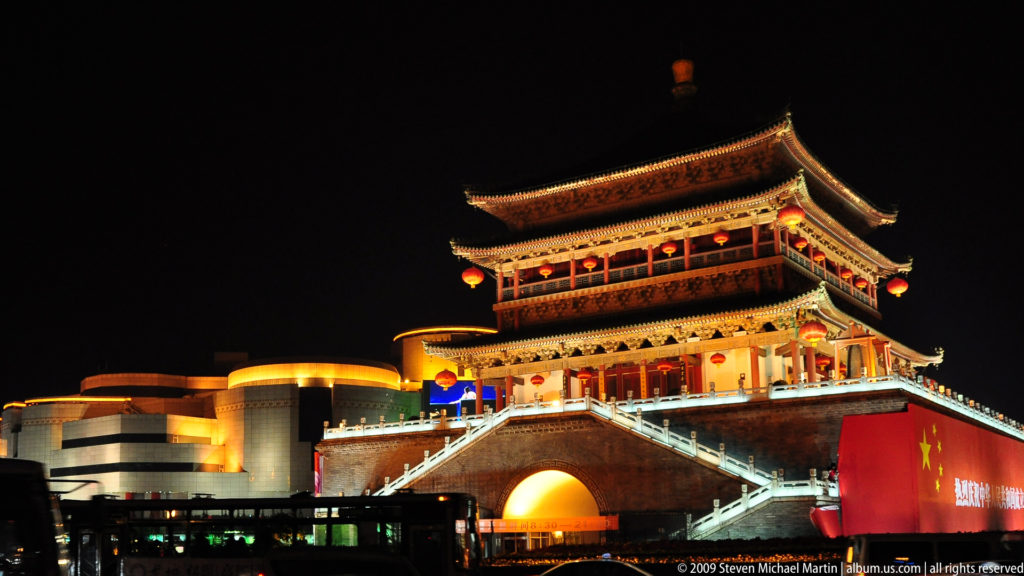 Front view of the illuminated Drum Tower of Xi’an at night, showing its tiered timber pavilion on a large brick platform with a central arched passage and symmetrical stairways.