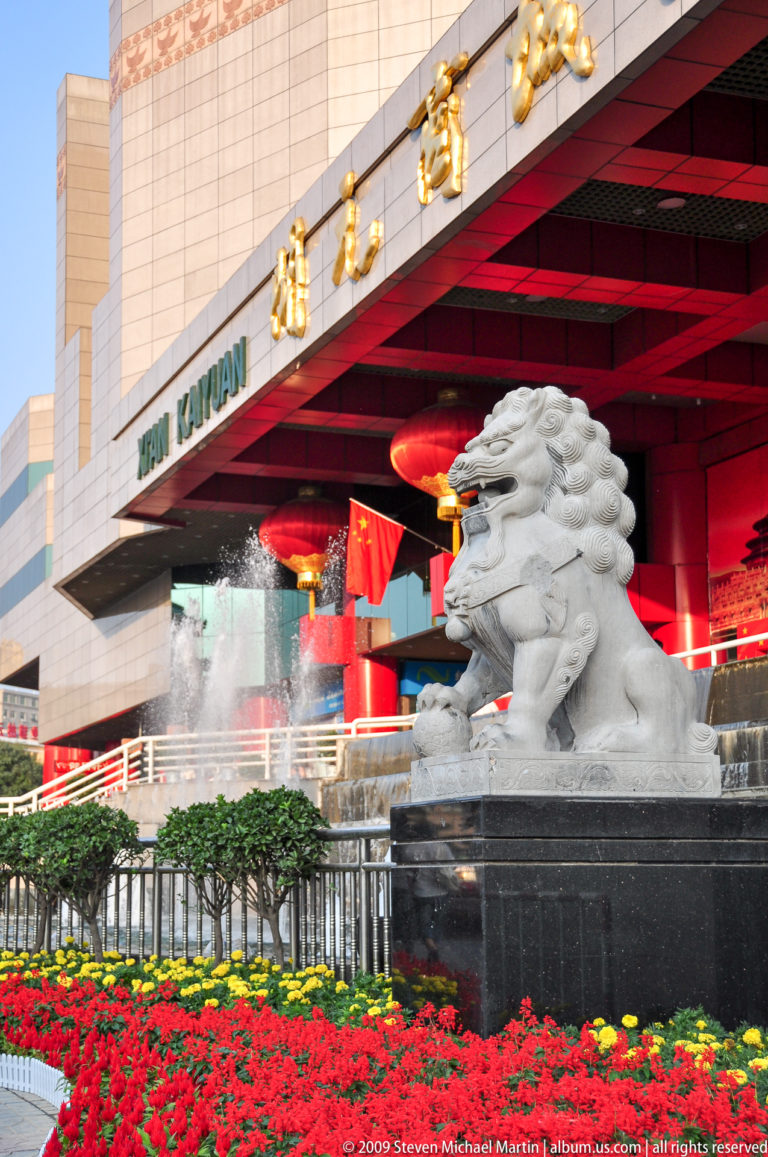 Large carved stone guardian lion with raised forepaw beside fountains, red columns, red lanterns, and Chinese national flags at a modern building entrance in Xi’an during a 2009 celebration.
