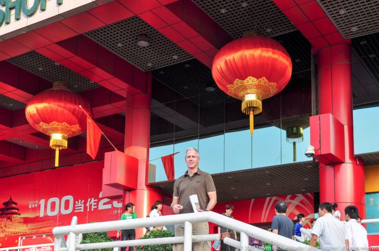 Male traveler standing behind a white railing at a modern entrance decorated with large red lanterns, Chinese flags, and 60th-anniversary graphics for the People’s Republic of China in 2009.