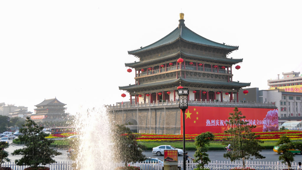 Bell Tower of Xi’an on a high masonry base at a busy city-center intersection, seen across fountains, flowers, and a red 60th-anniversary banner for the People’s Republic of China.