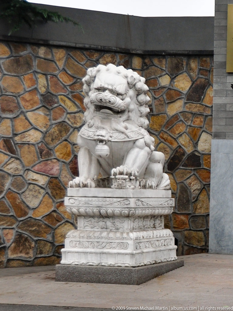 Pale stone guardian lion seated on an ornate carved pedestal before a wall of irregular brown stone.