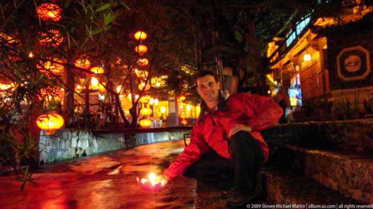 Male visitor crouching by a canal at night, extending his hand over the water with a glowing lotus lamp, with lantern-lit buildings and trees in the background.