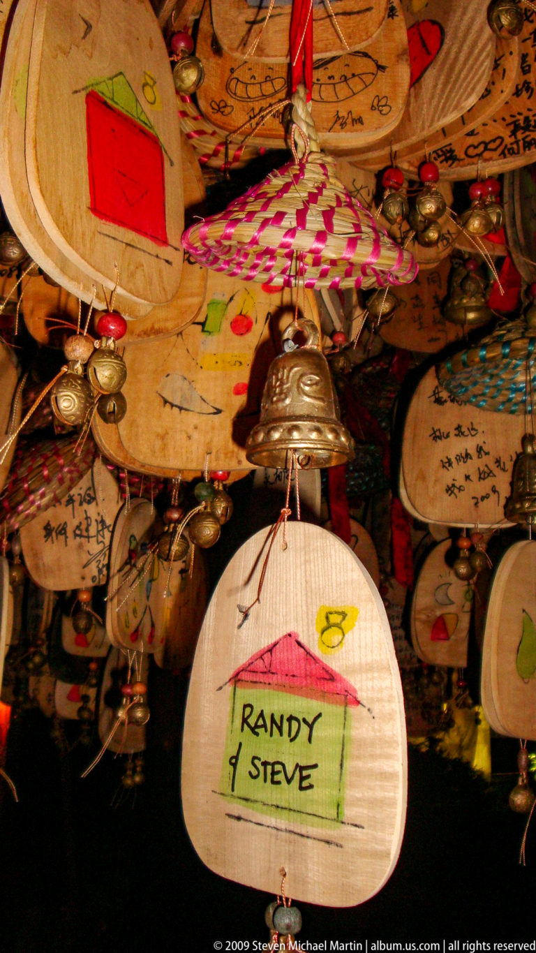 Close-up of metal souvenir bells hanging among inscribed wooden plaques with handwritten Chinese text and drawings in a market in Lijiang, Yunnan, China.