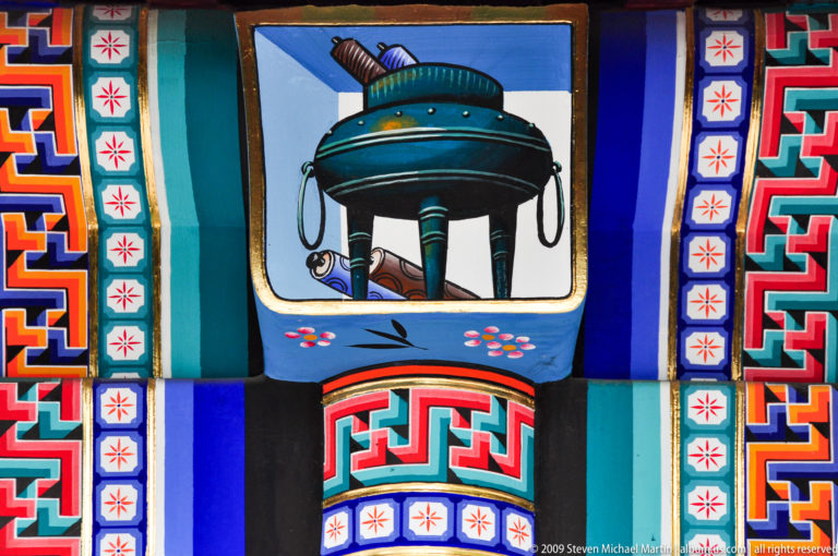 Colorful painted architectural panel with a stylized bronze ritual vessel motif framed by geometric red, blue, green, and white patterns.