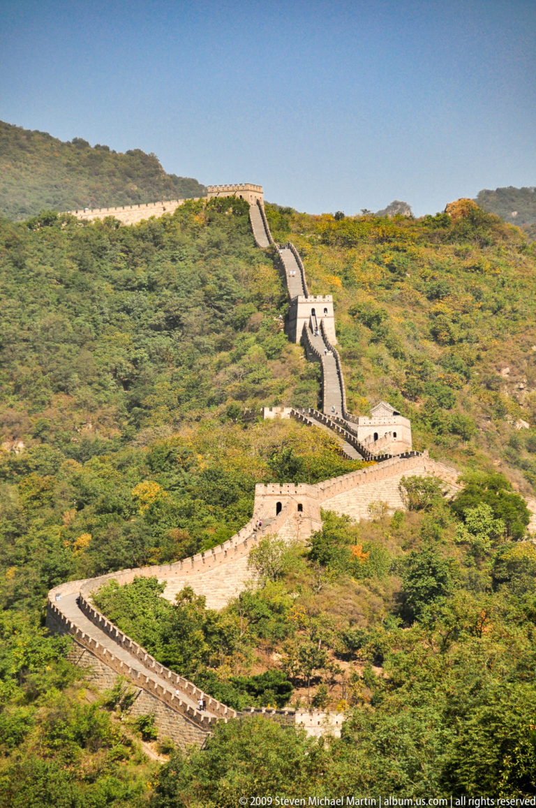 High-angle telephoto view of three closely spaced Great Wall watchtowers connected by very steep ramparts descending through green and gold autumn forest at Mutianyu.