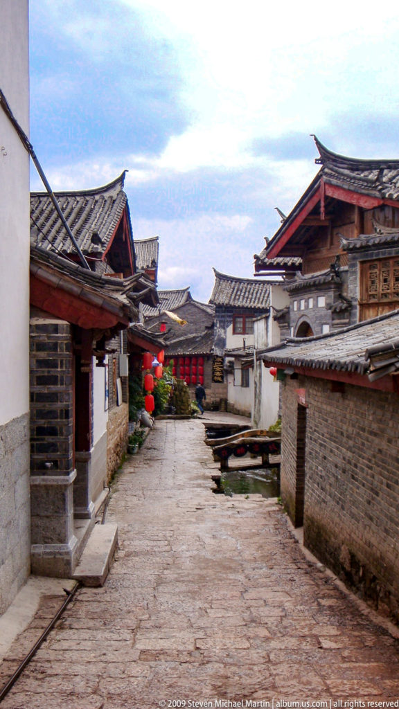 Narrow stone lane between traditional courtyard houses with overlapping grey tiled eaves, red lanterns, and a small water channel in Lijiang, Yunnan, China.