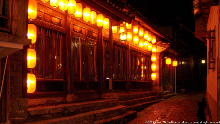 Traditional wooden building in Lijiang, China illuminated at night by rows of yellow lanterns under deep tiled eaves above dark timber doors and lattice windows along a stone street.