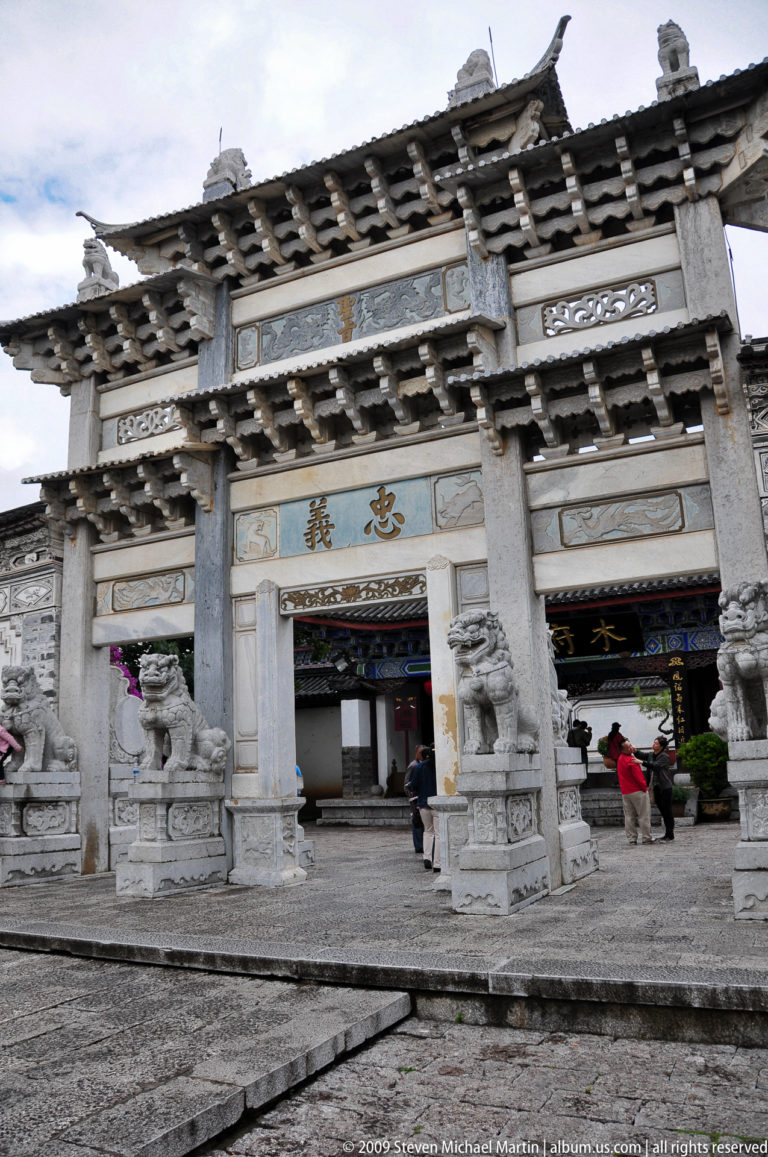 Ornate stone paifang-style gateway with layered bracketed roofs, large Chinese characters, and paired guardian lions in Lijiang, Yunnan, China.