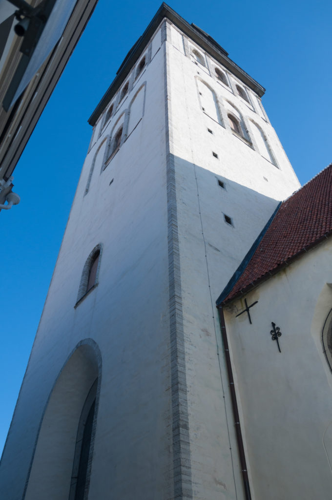Steep low-angle view up the white tower of St Nicholas Church beside a red tile roof and large pointed opening.