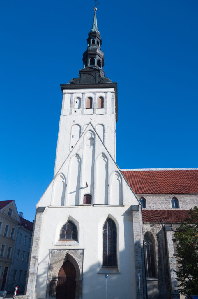 Frontal view of St Nicholas Church with pointed entrance, tall blind arches, massive white tower, dark lantern and red-roofed nave.