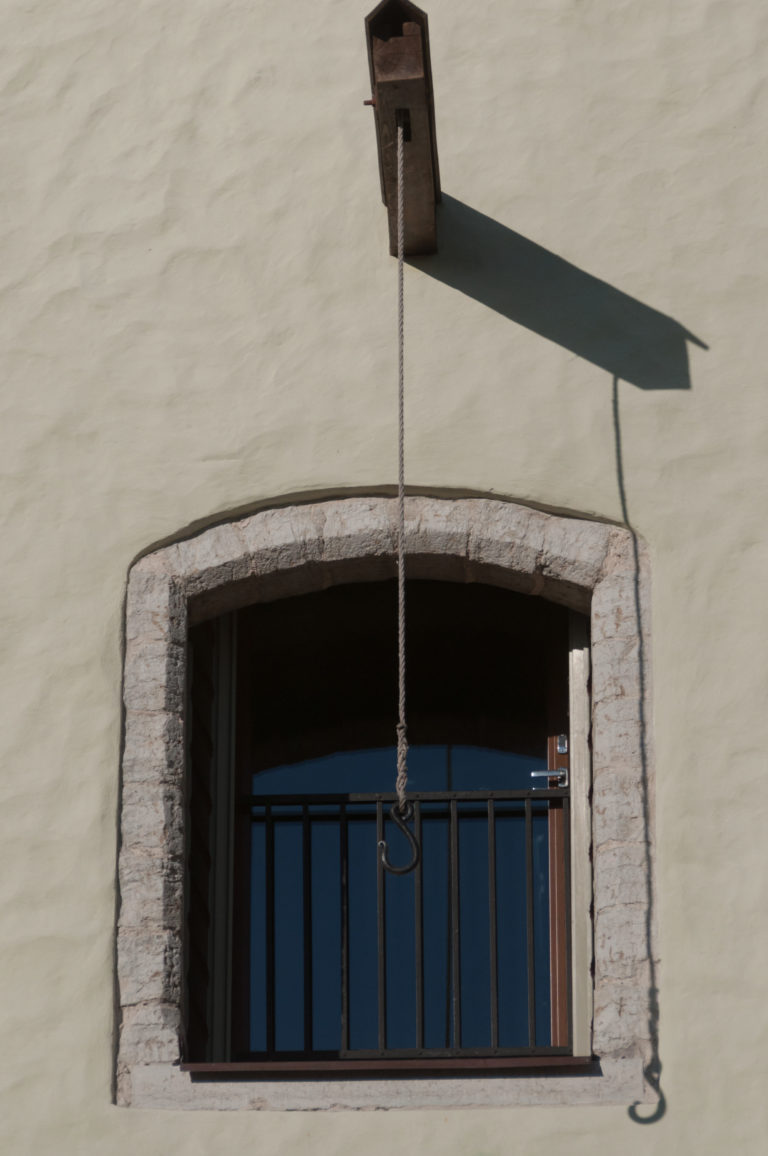 Barred stone-trimmed opening beneath a projecting wooden beam with a long hanging rope or chain ending in a metal hook.