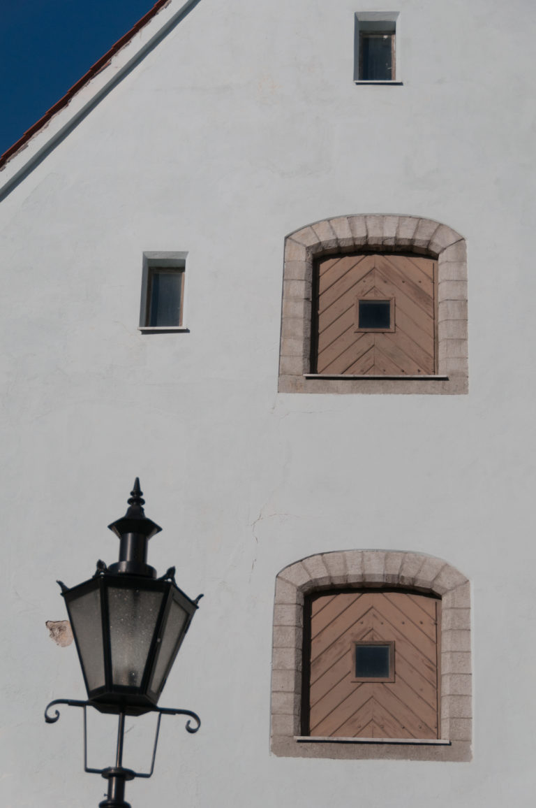 White church wall with two small stone-trimmed wooden shutters and a projecting black metal lantern at lower left.