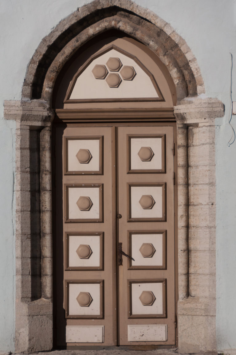 Paneled double wooden doors set within a pointed stone arch in the white exterior wall of St Nicholas Church.