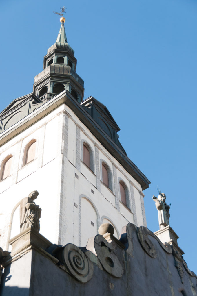 White upper tower of St Nicholas Church with arched openings and dark roof rising behind an ornate sculptural roofline.