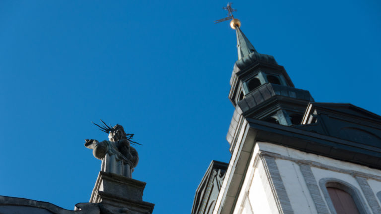 Upward view of St Nicholas Church tower and spire behind a rooftop sculptural figure against a clear blue sky.