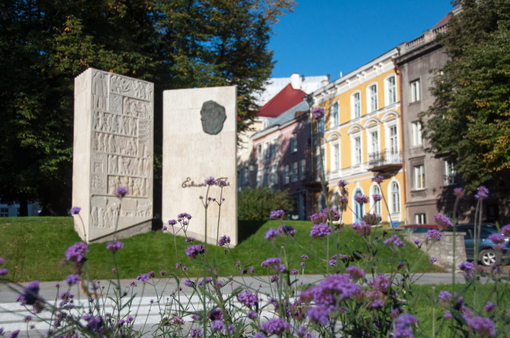Two upright stone memorial slabs beside purple flowers and colorful historic buildings in Tallinn’s Old Town.