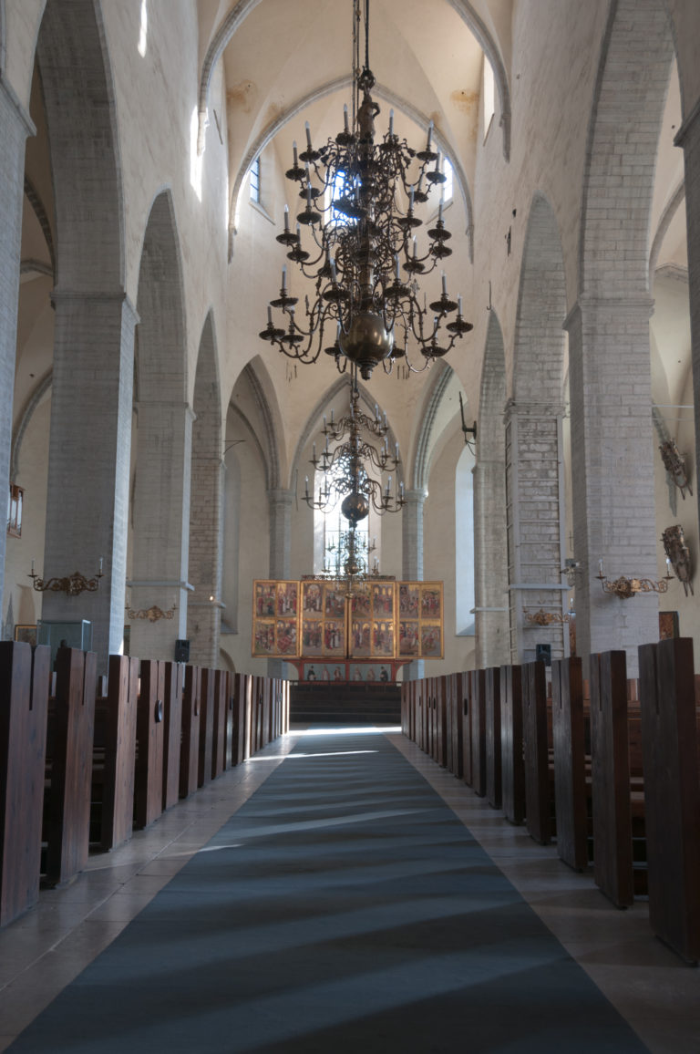 Low symmetrical view down the nave toward the high altar, with wooden seating, pale brick arcades and a chandelier centered overhead.