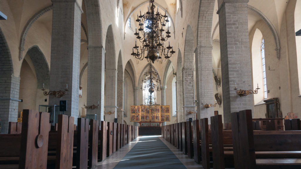 Oblique view through the nave with a large chandelier, wooden pews, pointed arches and the high-altar retable in the distance.