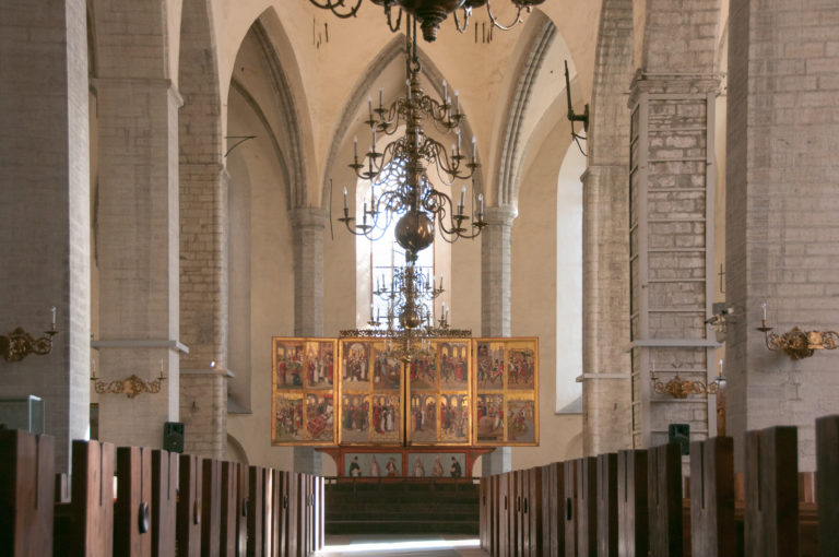 Central nave view between wooden pews toward the high-altar retable, with pale brick arches and large hanging chandeliers overhead.