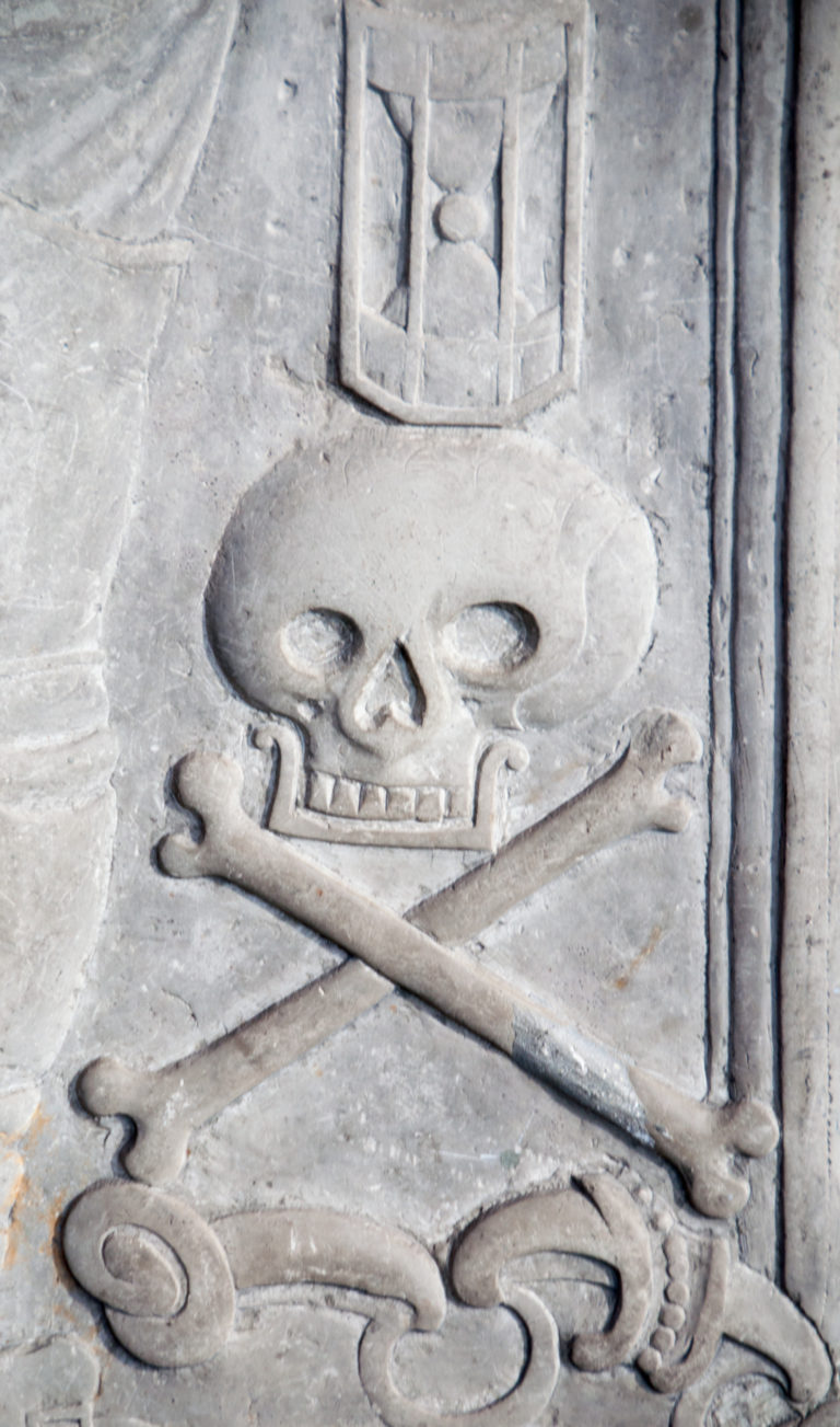 Close view of a carved skull and crossed bones in low relief on a weathered stone grave monument.