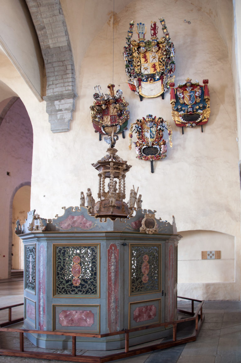 Painted enclosed church furnishing with dark lattice panels, red marbling, metal ornament and several colorful heraldic shields mounted above.