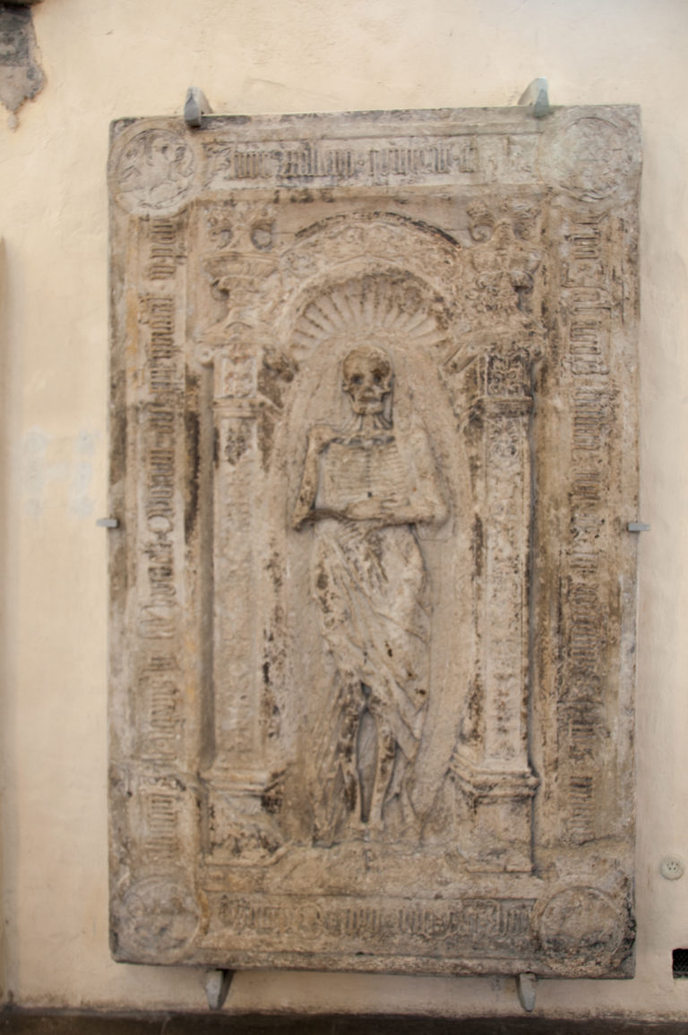 Weathered rectangular stone grave slab with a standing figure in shallow relief and an inscription running around the border.