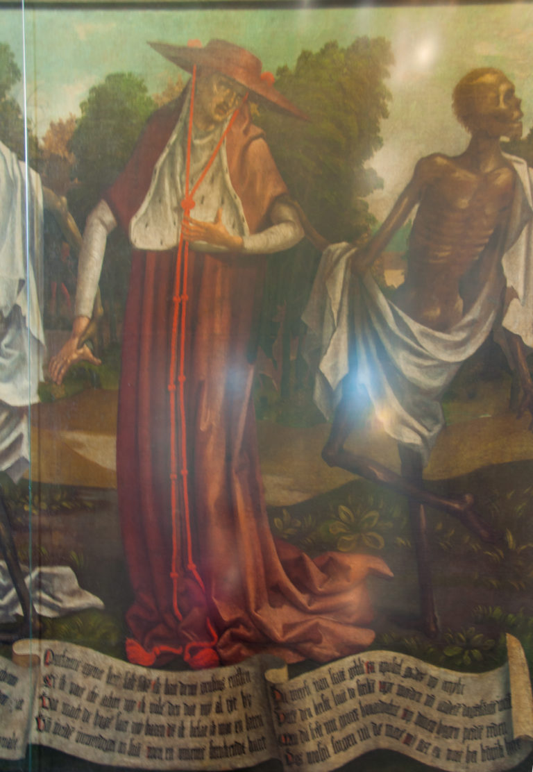 Close detail of the Dance of Death showing a red-robed courtly figure beside a skeleton above a scrolling Low German inscription.