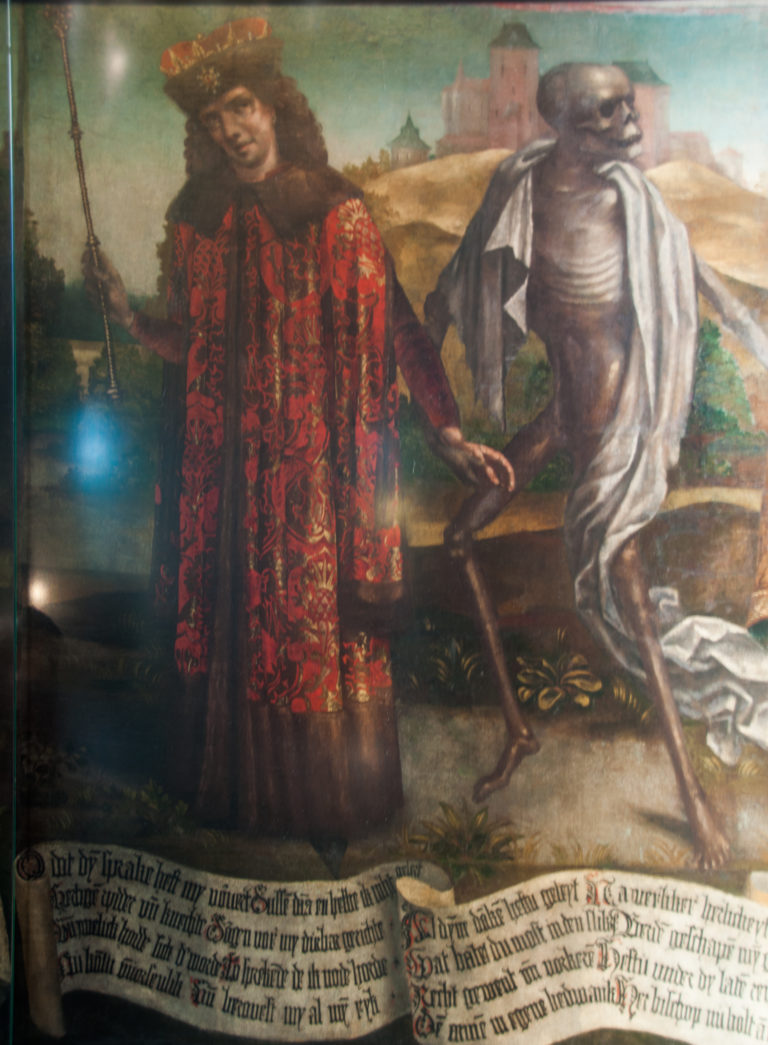 Detail of the Dance of Death with a richly dressed figure beside a shrouded skeleton and a band of Low German text below.