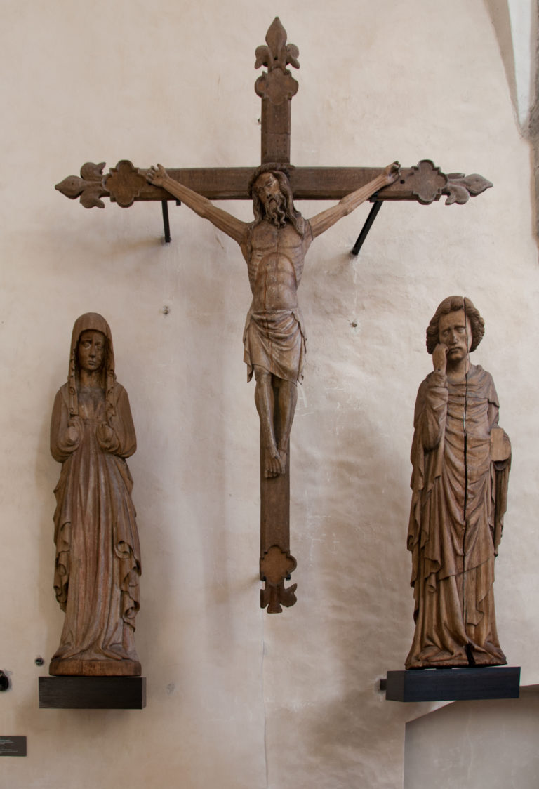 Carved Crucifixion group with Christ on the cross flanked by the Virgin Mary and St John against a pale wall.