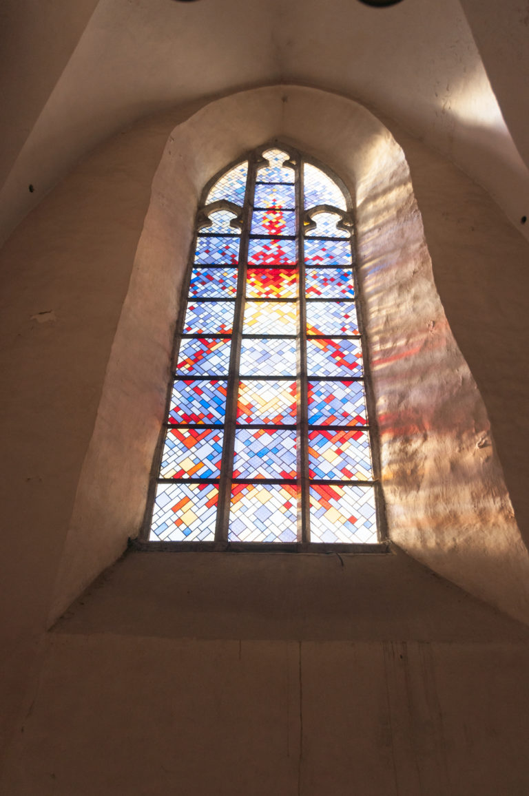 Upward view of a pointed stained-glass window with geometric colored panes and sunlight reflected on the surrounding plaster.