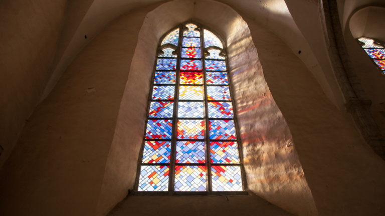 Tall pointed stained-glass window with red, blue and yellow geometric panes casting colored light across the deep wall reveal.